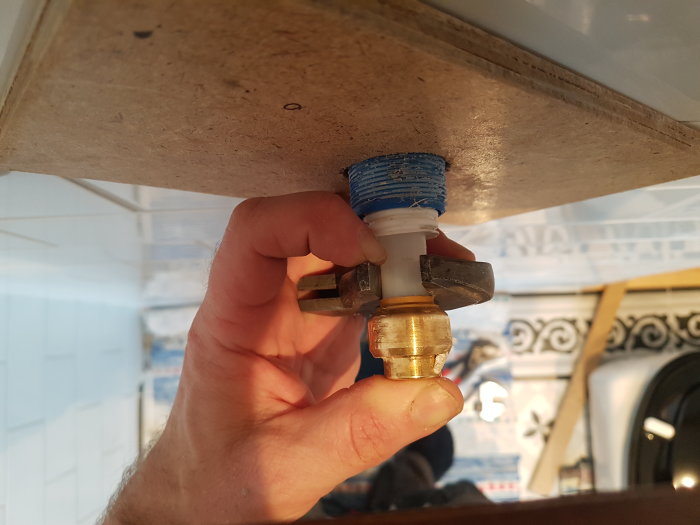 Hand håller en skiftnyckel som pressar en brun plastring i en mässingspropp under en vattentank.