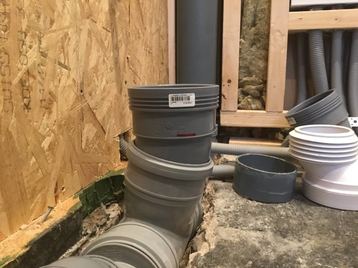 VVS-installation med excentrisk WC-stos och böjda rör nära oslipade betonggolv och trävägg.