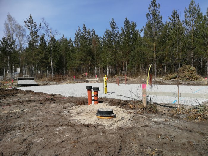 Rör sticker upp från marken vid en husgrund under konstruktion, omgivna av skog och grus.