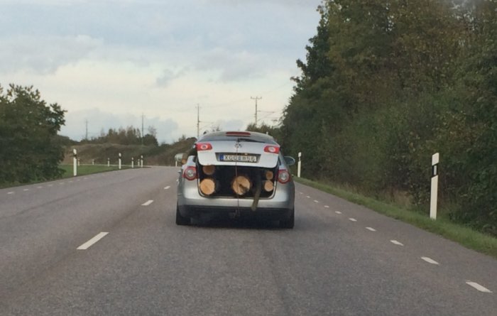 Bil överlastad med stora trädstammar som sticker ut bakifrån bagageluckan på en landsväg.