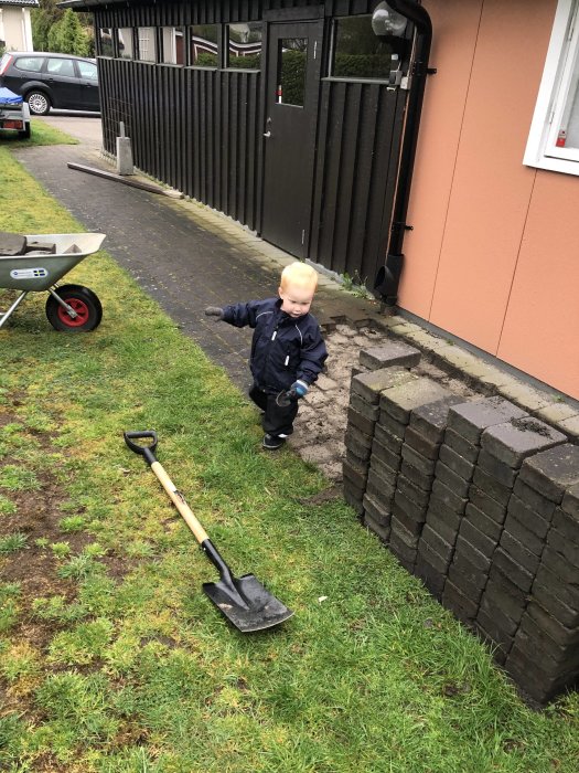 Ett litet barn står bredvid en hög av borttagen marksten och en spade, vid sidan av ett garage.