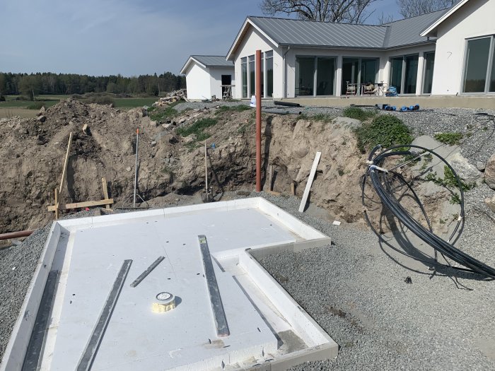 Grundläggning för ett pumphus vid en slänt med grus, rörledningar och isolerad betongplatta framför ett hus.