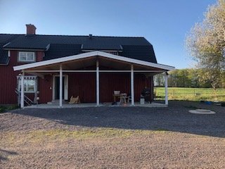 En röd villa med svart tak och tillbyggt överbyggt uterum, omgiven av en gruslagd tomt och gröna fält.