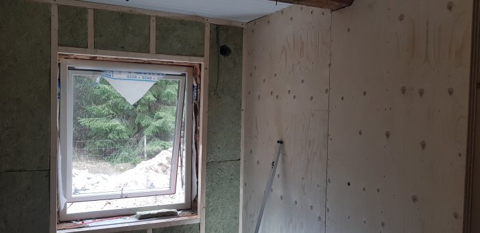 Inre hörn av ett rum under renovering med isolering och plywoodväggar, utsikt genom fönstret till träd och grushög.