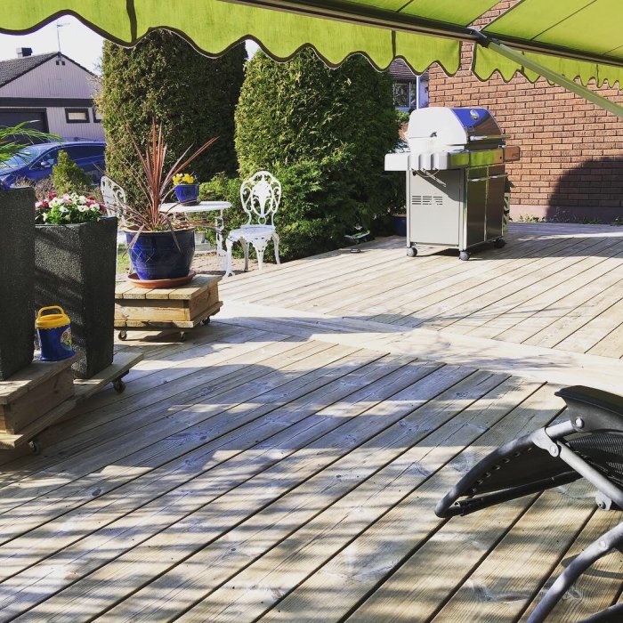 Nybyggd träterrass med utemöbler och grill, planteringar och en kruka på en pall under ett grönt markistak.