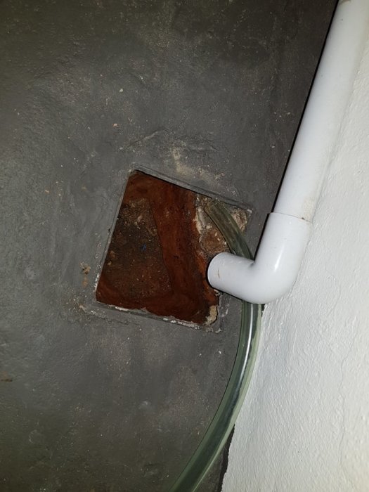 Gammalt avloppshål i betonggolv med synlig brunnskant och anslutande vita och gröna rör.