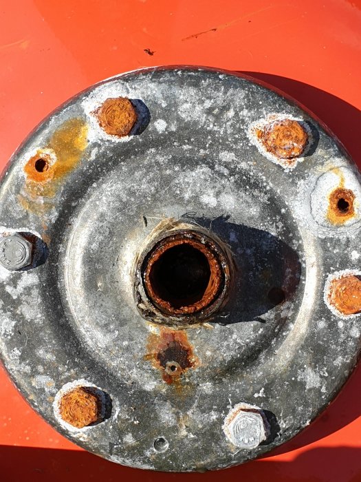 Rostig metallplatta med ett centralt hål, omgivet av rostiga bultar och korrosion, visar tydliga tecken på slitage och igensättning.