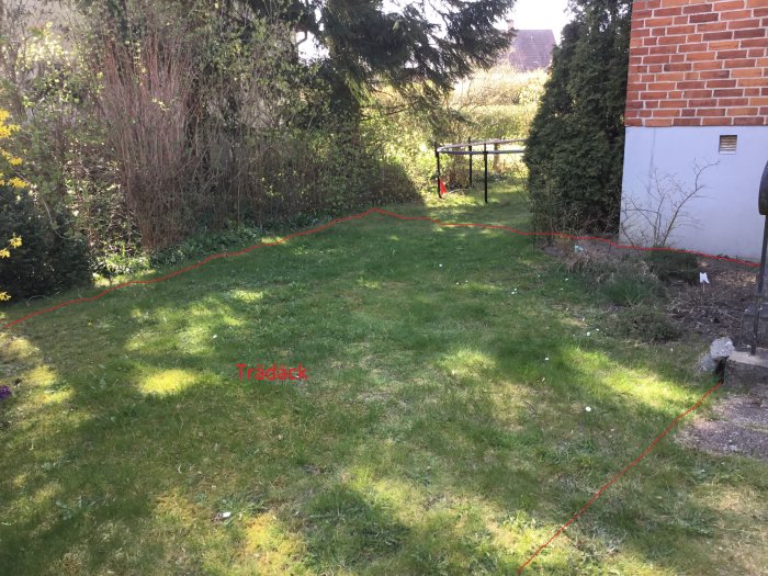 Trädgård vid hus med utmarkerat område för planerat trädäck nära en trappa och vegetation.