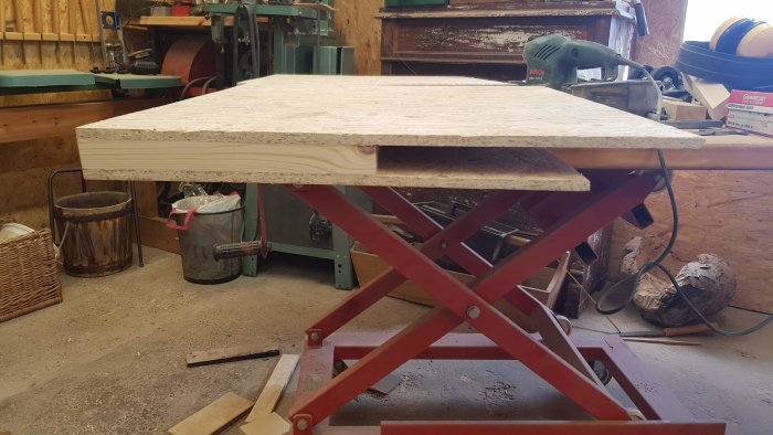 Handgjort skrivbord av reglar och OSB-skivor med korkmatta på ytan, placerat på rött lyftbord i en verkstad.