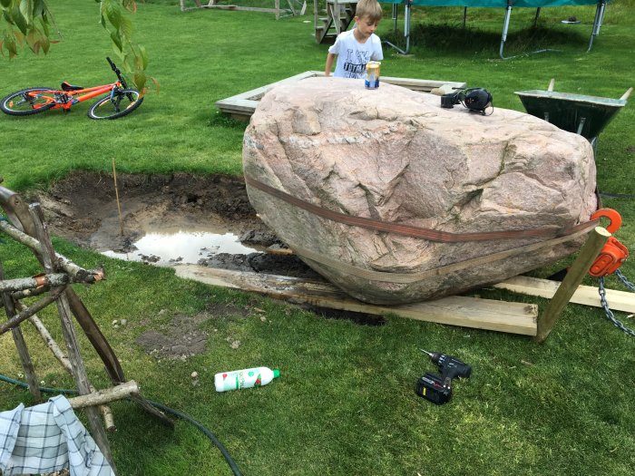 Stor sten säkrad med remmar ovanför grävd grop, barn och verktyg i närheten.