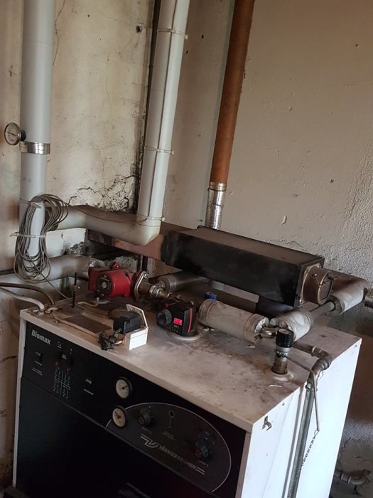 En pannrumsmiljö med en äldre värmepanna, diverse rörledningar och elkablar.