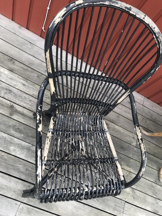 Svart stol med nätliknande rygg och sits, flagnande färg, på ett grått trägolv med röd vägg i bakgrunden.