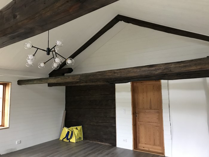 Renoverat rum nästan klart med vita väggar, träbjälkar, och modern taklampa.