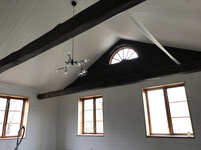 Inre del av ett rum under renovering med vitmålade väggar, mörka träbjälkar och flera fönster, inklusive ett halvmånefönster.