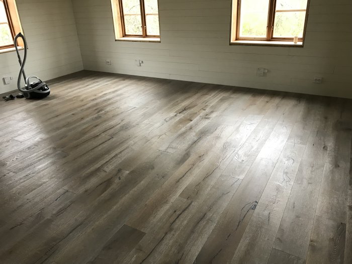 Oinreddt rum med nyinstallerat ljusgrått trägolv, målade väggar och fönster samt en dammsugare i hörnet.