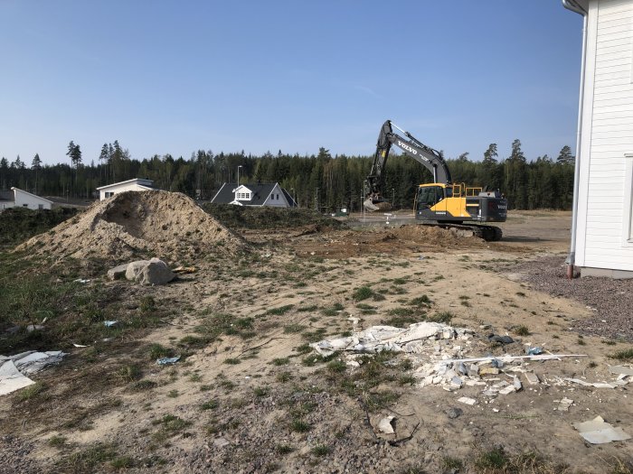 Avschaktad tomt med grushög, grävmaskin och nybyggda hus i bakgrunden.