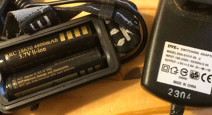 Bild på en pannlampa med märkta batterier bredvid en laddare med specifikationsetikett.