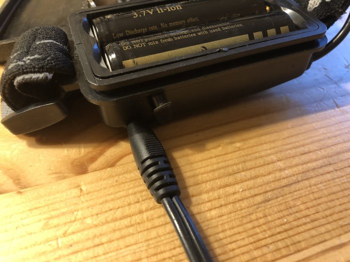 Svart laddningssladd ansluten till ett batteripack för pannlampa på en träyta.