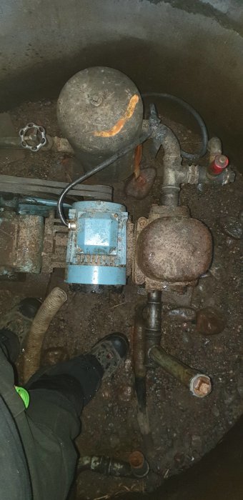 Gammal kolvpump med tätade anslutningar i en brunn, omgiven av lerig jord, verktyg och slangen synlig.