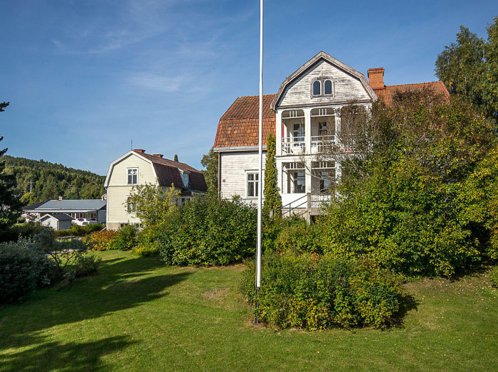 Traditionellt hus byggt 1916 med vit och röd tegel på taket, omgiven av grönt gräs och buskar under klar himmel.