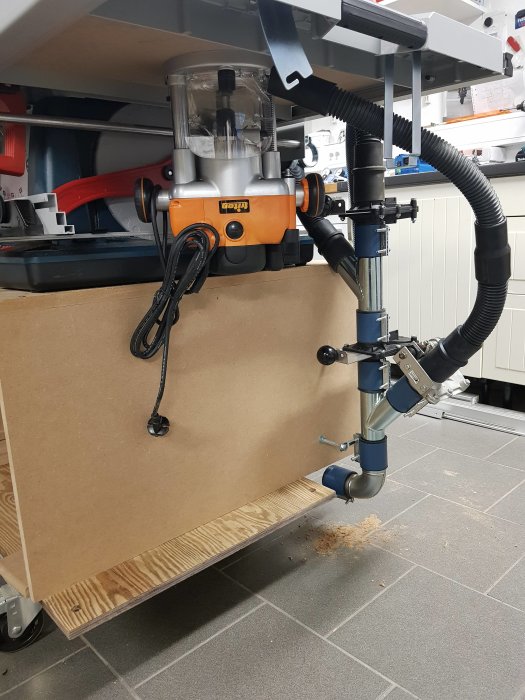 En fräsmaskin monterad under en arbetsbänk ansluten till ett dammutsugssystem med spånhög på golvet.