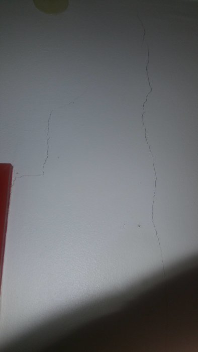 Stora sprickor i en ljus vägg som sträcker sig uppåt och runt ett hörn, indikerar möjlig sättningsskada.