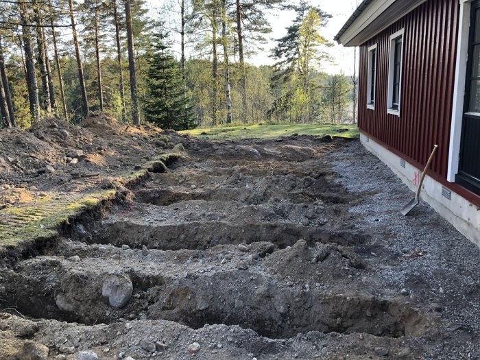 Uppgrävd mark för altanbygge nära ett rött hus med vita knutar och skog i bakgrunden.