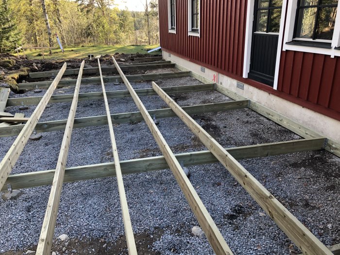 Regelverk till en altan under konstruktion intill ett rött hus med vita fönster, omgiven av natur.