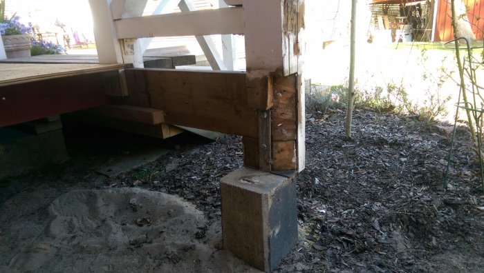 Verandans hörnstolpe vilar på en liten träkloss ovanpå en betongplint, intill ett nytt trädäck.