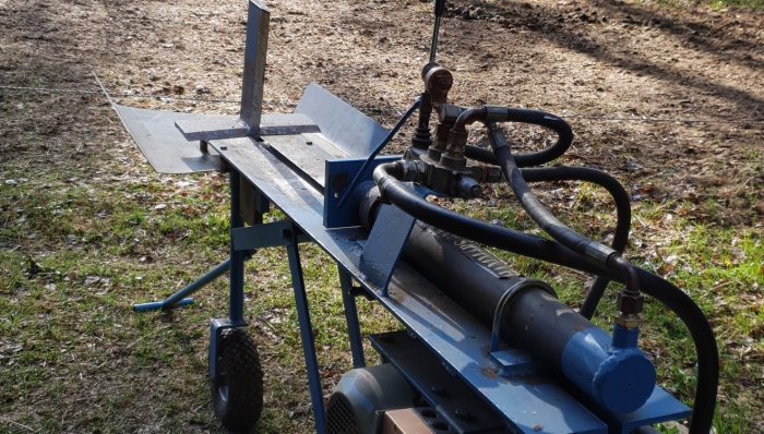 En väderbiten vedklyv utomhus med synliga hydraulslangar och kolven delvis utsatt, omgiven av gräs och löv.