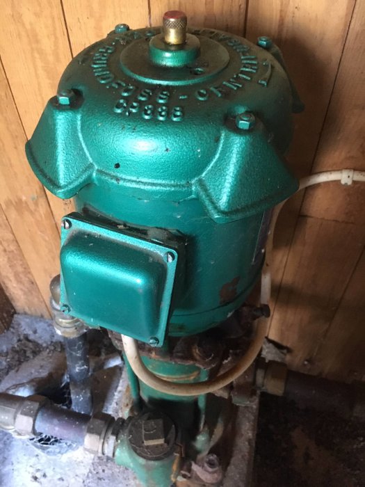 Gammal grön Grundfos vattenpump i ett torp, modell CP 898, med synliga rostiga röranslutningar och en smutsig bakgrund.