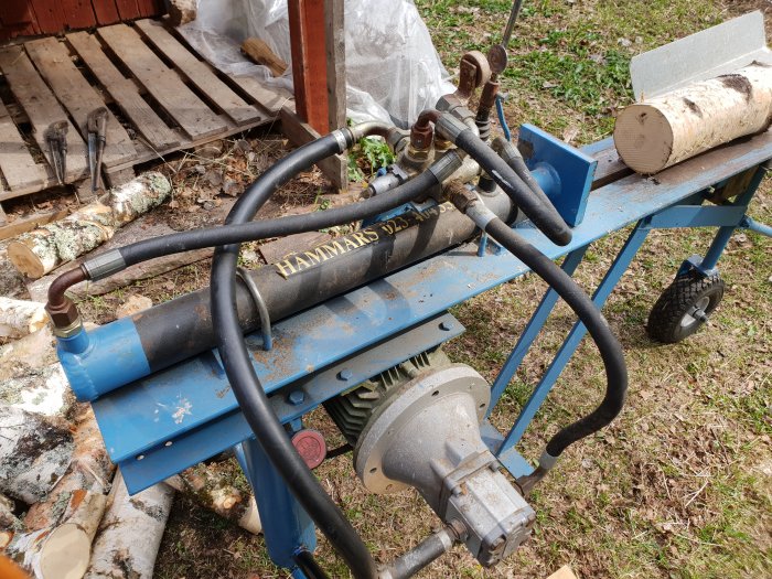 Hydraulisk vedklyv med flera svarta slangar och metallkomponenter på en blå vagn utomhus.