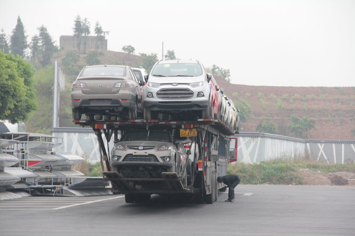 Lastbil transporterar en hög stapel av bilar på en väg i Kina, med en person som tittar på underfordonet.