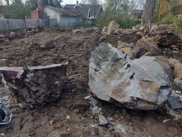 Två stora stenblock med spräckkil i en grävd jordlott omgivna av röjda stenar och byggnader.