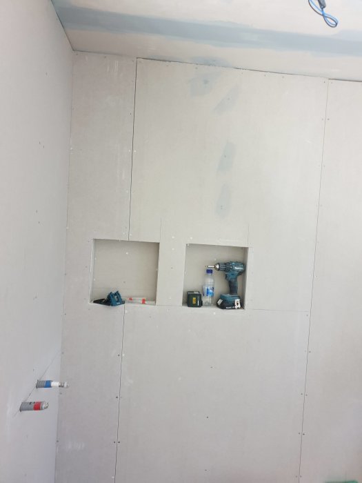 Vägg i pågående badrumsrenovering med gipsskivor, utskärningar för rör och el, samt en borr och verktyg.