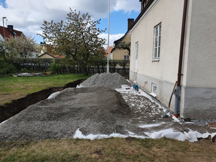 Högar av makadam vid husgrund för utjämningsarbete med grävd dikeskant i bakgrunden.