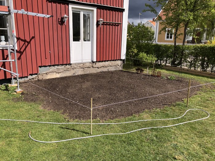 Rödfärgat hus med borttagen uteplats framför fasaden, redo för renovering, med bar jord och gräsmatta runtomkring.