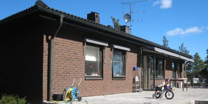 Ett enplans tegelhus med terrass, fönster och en skorsten, plus en barnvagn och cykel i förgrunden.