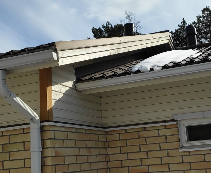 Hörn av ett hus med snötäckt tak, ventilationsrör, och synlig husisolering under en solig dag.