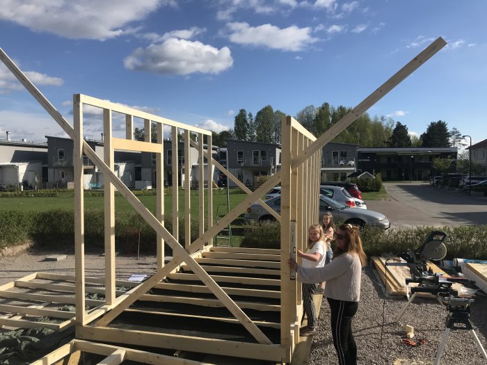 Familj hjälper till att resa en trästomme för en vägg i ett byggprojekt medan en cirkelsåg och trämaterial syns i förgrunden.