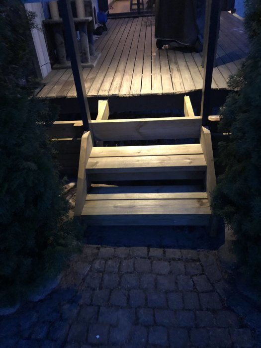 Trappa i vitt trä leder upp till en träaltan med belysning ovanför, framför ligger en yta med gatsten, tagen i skymning.