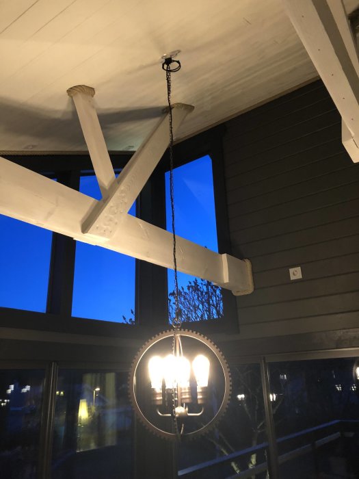 Taklampa med tända glödlampor hänger i uterum med mörk vägg och synliga takbjälkar.
