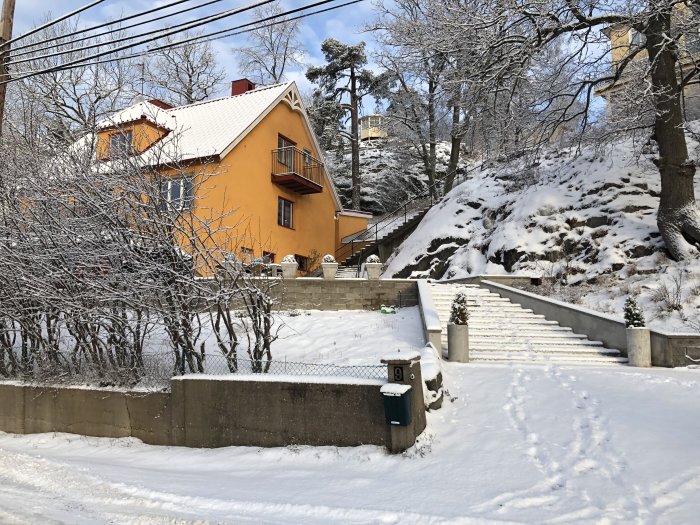 Vinterbild på en lutande mur framför ett gult hus med snötäckt trädgård och trapp.