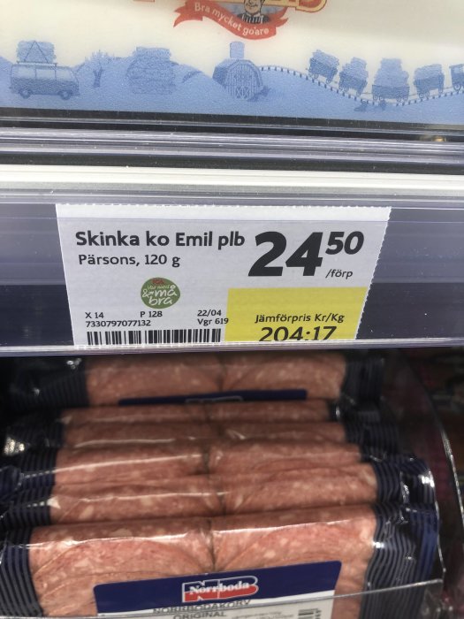 Prisetikett på "Skinka ko Emil" ovanför förpackade korvar i butikshylla, ironisk anspelning på genmodifierad mat.