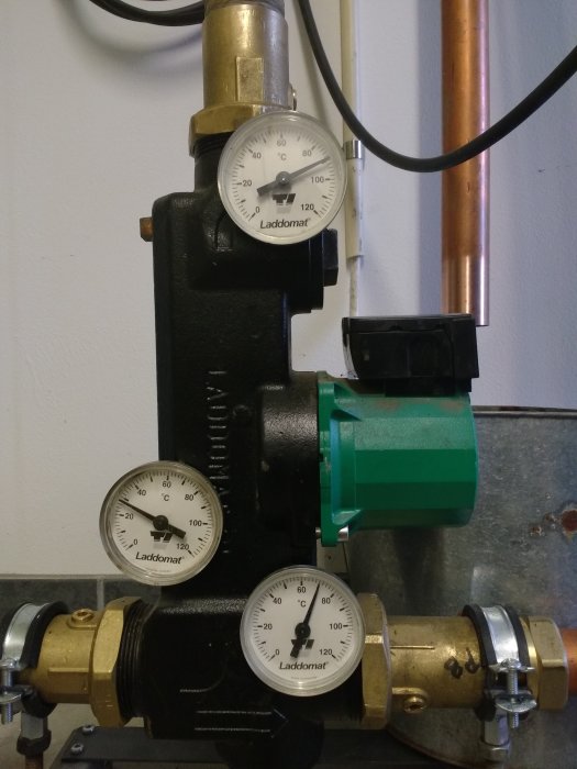 Laddomat för värmesystem med tre termometrar och en grön pump mot en vit vägg.