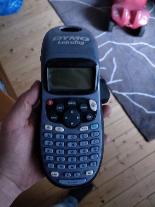 Hand som håller en DYMO LetraTag etikettskrivare med trätak och en leksaksbil i bakgrunden.