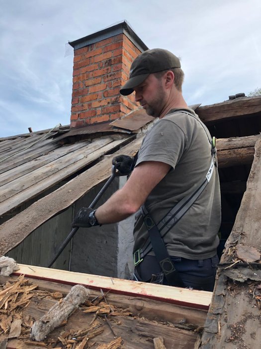 Person i arbetskläder renoverar tak nära skorsten, justerar trämaterial mot vägg.