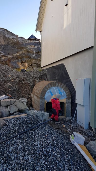 Nymurad ingång till en jordkällare med dräneringsplast och omgivande grus, stenar och barn framför.