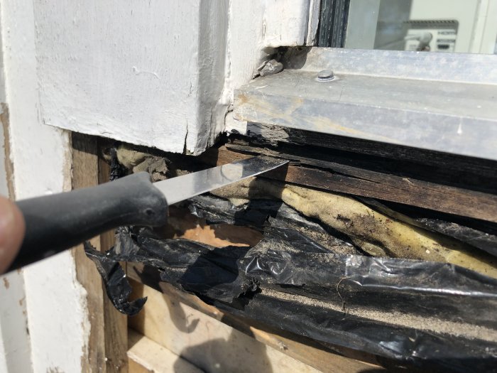 Skadad fönsterkarm med synlig röta och svart plast, kniv används för att skrapa bort dåliga delar.
