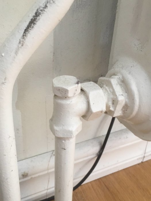 Närbild av vitmålad radiatoranslutning och justerventil intill en vägg och golvlister.